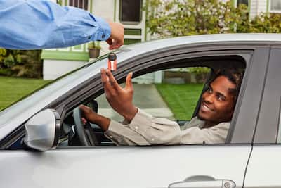 Young man in the drivers seat getting the keys to the vehicle from someone standing outside the car.